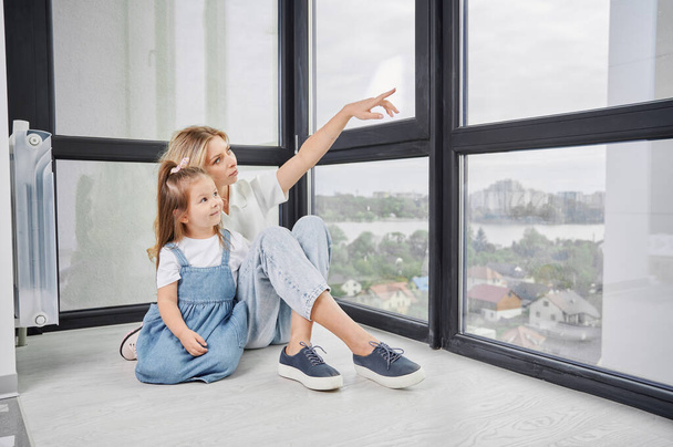 Nainen ja pikkutyttö istuvat lattialla ja katsovat ja osoittavat ikkunaa perheen uudessa kodissa. Äiti ja lapsi nauttivat näköalasta panoraamanäkymästä hiljattain rakennetussa huoneistossa. - Valokuva, kuva