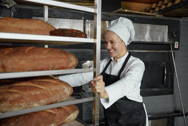 Experienced woman baker smiling happily pushing metal rack with fresh baked bread baguettes working in bakehouse - Photo, Image