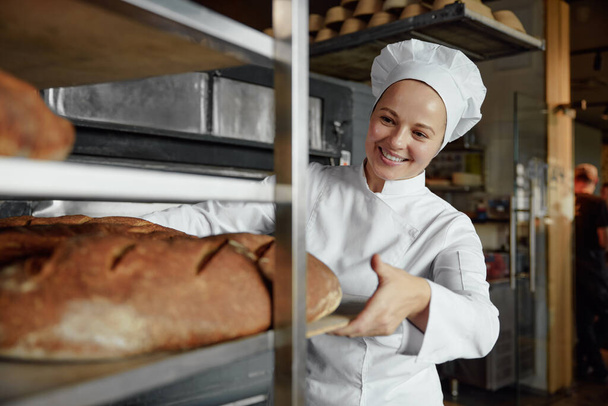 Kobieta piekarz uśmiecha się szczęśliwie pchając drewniany stojak ze świeżo upieczonymi bagietkami chlebowymi pracującymi w piekarni - Zdjęcie, obraz