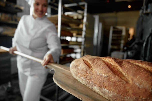 Lähikuva midsection näkymä juuri leivottu käsityöläinen leipää puinen lapio naisten leipuri käsissä. Leipomo valmistus käsite - Valokuva, kuva