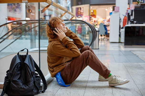 Menino adolescente relaxante no shopping sentado no chão ouvindo música e desfrutando de tempo livre sozinho. Conceito de emoção despreocupada - Foto, Imagem