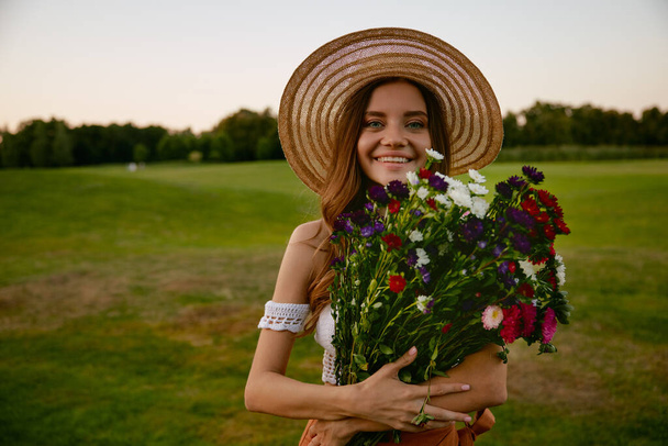 Портрет лица красивой хипстерки, держащей букет диких цветов, стоящих над зеленым загородным полем. Женственность, природная красота и невинность - Фото, изображение