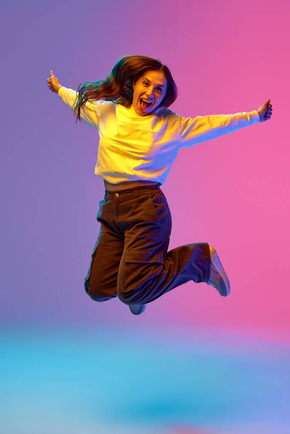 Full-length portrait of young positive woman jumping of joy raising hands against gradient purple-blue-pink background. Concept of beauty, youth, human emotions, self-expression, fashion and style. - Photo, Image
