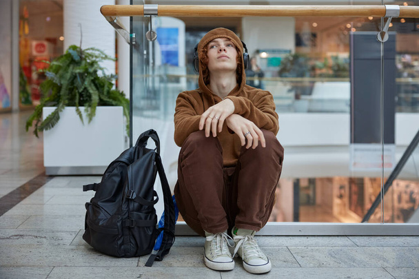 Upset teenage boy in headphones sitting nearby moving staircase at shopping mall. Depressed serious guy listening to music feeling negative emotion - Photo, Image