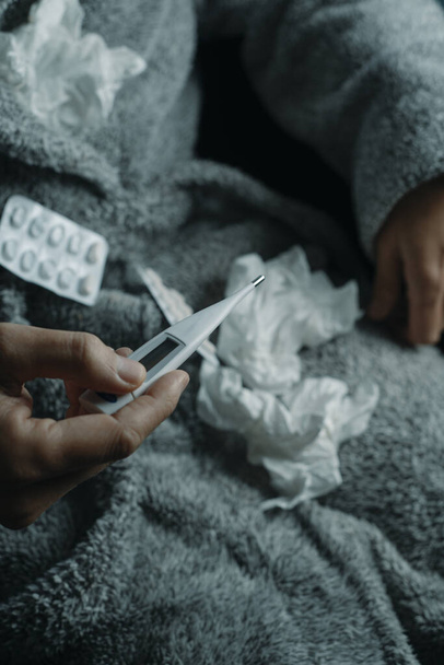 um homem doente em casa, vestindo um roupão cinza macio, cercado por tecidos usados, está prestes a medir sua temperatura com um termômetro - Foto, Imagem