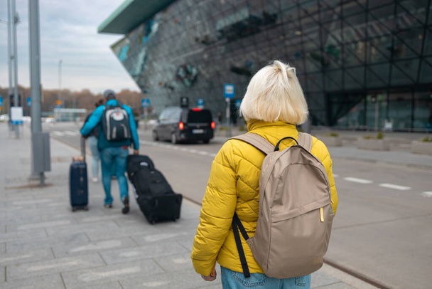 Счастливая привлекательная 60-летняя пожилая женщина шла с чемоданом на современной остановке общественного транспорта рядом с современным терминалом аэропорта. Отдых, путешествия и активный образ жизни  - Фото, изображение
