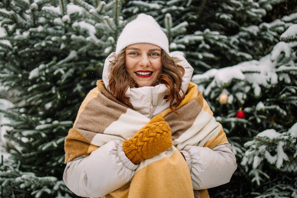 Portrait of smiling happy caucasian woman in hat with mittens in winter coat laughing, tossing snow up outside in the city park in winter. High quality photo - Photo, Image