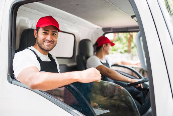 赤い帽子をかぶったアシスタントと幸せなプロのトラック運転手,微笑んで,小包を配達する前にトラックの窓からカメラを見ています. トラックドライバーと配送サービスのコンセプト. - 写真・画像