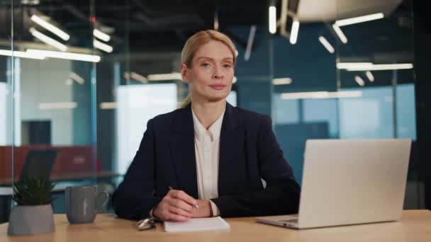 Portrait d'une femme d'affaires confiante et sérieuse assise au bureau, croisant les bras, regardant la caméra avec un sourire parfait. Une entrepreneure indépendante termine son travail dans un bureau en verre moderne. - Séquence, vidéo