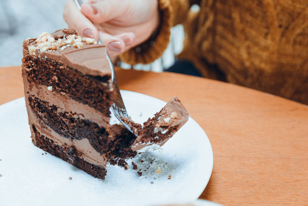 hand of unrecognizable caucasian woman cutting with fork slice of chocolate cake served on white porcelain plate on wooden table, pastry concept for social media, copy space - Photo, Image