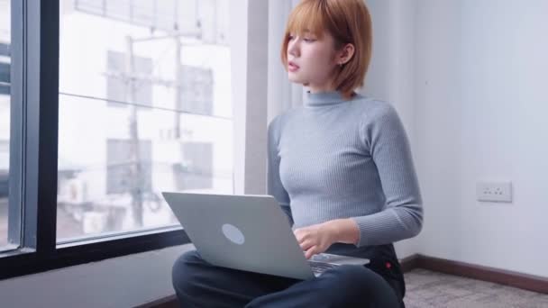Уверенная азиатская деловая женщина, сидящая у стеклянного окна в офисе и усердно работающая над своим ноутбуком, демонстрируя целенаправленное и решительное отношение. Высококачественные 4k кадры - Кадры, видео