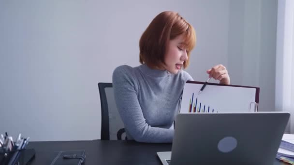 Una mujer de negocios asiática confiada, presentando diligentemente el desempeño trimestral de la compañía a través de una video llamada decidida en su computadora portátil en su espacio de trabajo bien organizado. - Metraje, vídeo