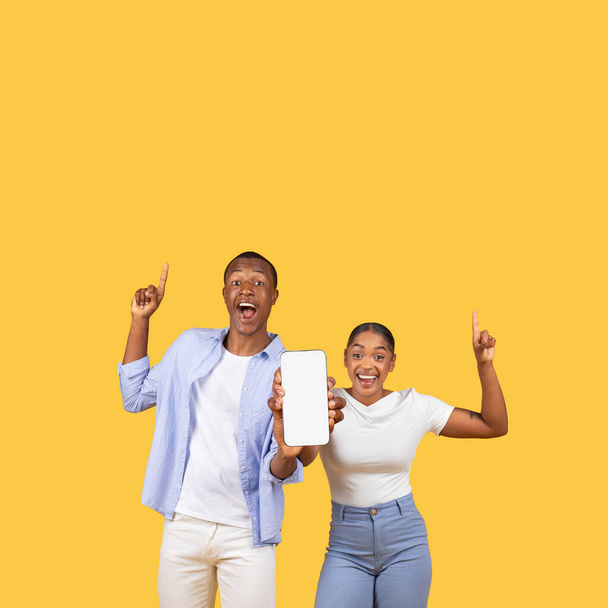 Joyful black couple pointing upwards and presenting smartphone screen with blank space for content, embodying excitement and surprise, against yellow background, mockup - Zdjęcie, obraz