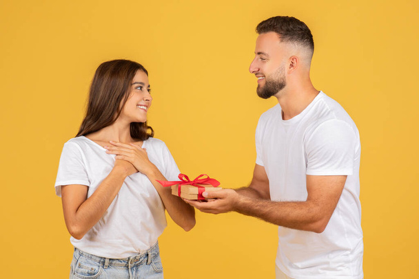 A joyous young caucasian woman looks at a smiling man who is giving her a small gift box with a red ribbon, representing a gesture of affection and surprise on a yellow background - Foto, Imagen