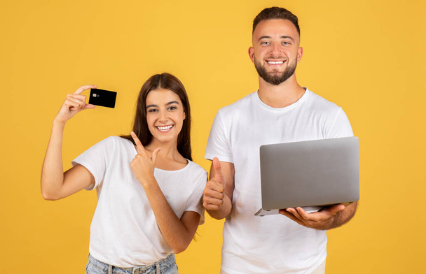 Confident young caucasian woman holding up a credit card with a thumbs-up as a pleased man holds a laptop, both endorsing online financial transactions on a yellow backdrop - Foto, imagen