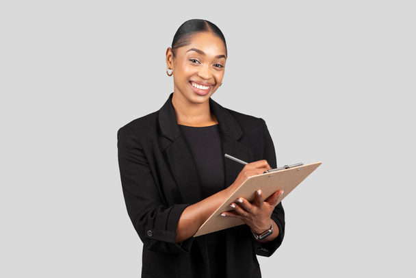 Positive pretty young african american lady secretary, manager in formal suit make notes at clipboard, isolated on gray studio background. Business, work and study, ad and offer - Foto, Imagen