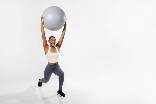 Young black woman in fitwear lifting stability ball while exercising, smiling confidently, over white studio background. Strength and wellness, muscular training. Empty space - Foto, Imagem