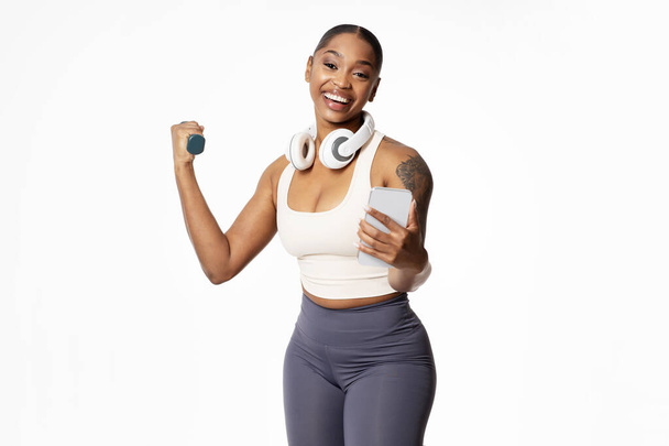 Cheerful black athletic lady with earphones headset holds smartphone while flexing arm muscles with dumbbell, standing in studio over white background. Fitness and strength workout app - Photo, image