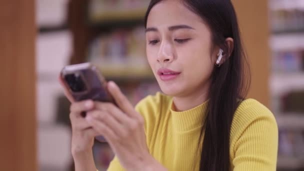 nice Asian student woman sitting at table and using cellphone or smartphone texting chatting social media or listening song while doing homework in library or reading room. High quality 4k footage - Footage, Video