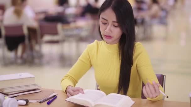 Koncentrált fiatal ázsiai lány diák sárga ruhában könyveket olvas kutatás tanulmány asztalra a főiskolai közkönyvtárban. Kiváló minőségű 4k felvételek - Felvétel, videó