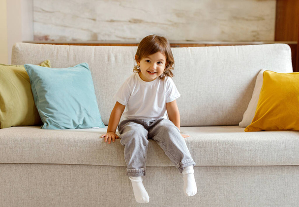 Cheerful little girl lounging on sofa at home, grinning brightly at camera, cute preschooler female child relaxing on on comfortable couch in living room, capturing the essence of youthful exuberance - Photo, image