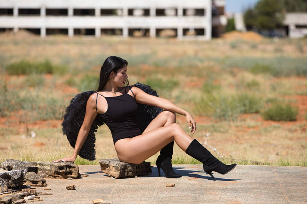 Eine junge, schöne, dunkelhaarige lateinische Frau mit schwarzen Flügeln steht inmitten der Ruinen einer von Krieg und Apokalypse zerstörten Stadt und blickt sich mit Traurigkeit und Trauer um. - Foto, Bild