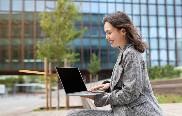 Young businesswoman browsing web on laptop sitting outdoors, representing IT and software career in urban street setting. Side view of lady websurfing on computer with empty screen mockup - Foto, Imagem