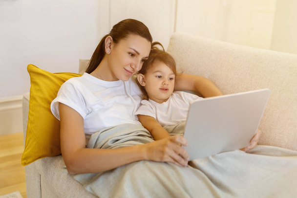 Mother and her little daughter with digital tablet relaxing under blanket on sofa, mom and preschool female child exploring online content together, watching cartoons or browsing internet - Foto, Imagen