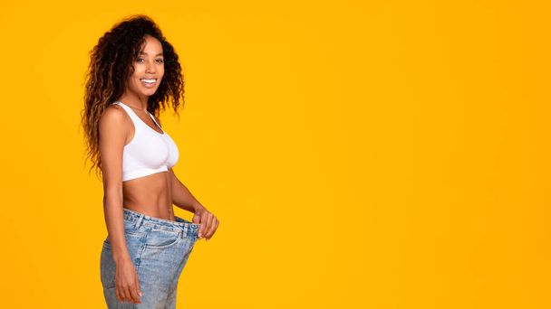 Perfect slim body. Smiling black woman wearing too large jeans proudly showing slimmed perfect body on yellow studio background. Healthy weight loss. Panorama, copy space - Photo, image