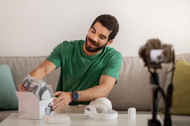 Delighted middle eastern young man in a green shirt engagingly unpacks new headphones from a box, sharing his excitement during a gadget review session for his vlog audience - Foto, imagen