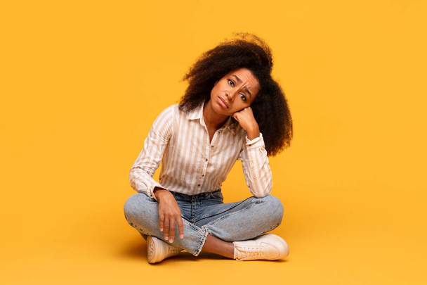 Contemplative young black woman with voluminous curly hair sits cross-legged on floor, hand resting on her cheek, deep in thought against vivid yellow background - Фото, изображение