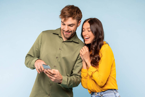 Delighted couple closely sharing joyful moment looking at smartphone, their expressions filled with laughter and happiness, against calming blue background - Fotó, kép