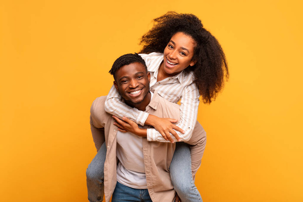 Cheerful young black man giving a piggyback ride to an excited woman, both sharing genuine smile, set against warm yellow background radiating happiness - Photo, image