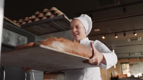 Woman baker smiling happily pushing wooden rack with fresh baked bread baguettes working in bakehouse - Footage, Video