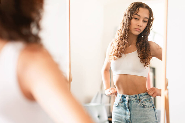 Unhappy teen girl looking critically at her thin waist in the mirror, suffering from discontent with her body and lack of self love and acceptance, standing at home. Anorexia concept - Foto, imagen