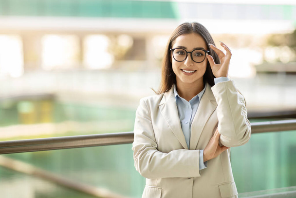Professional young pretty caucasian woman manager, boss with glasses smiling confidently, dressed in a light beige blazer, standing in a modern office environment in city - Foto, imagen