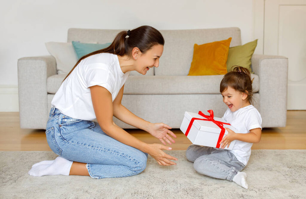 Excited little daughter receiving gift from mom at home, happy cute preschool female child getting birthday present box with red ribbon, sitting with mommy on floor in living room interior - Foto, afbeelding