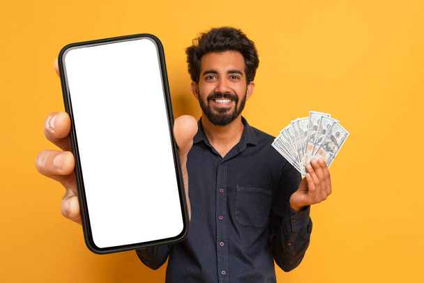 Cool Indian Man Holding Dollar Cash And Phone With White Empty Screen In Hands And Smiling, Happy Guy Showing Money Banknotes, Yellow Background. Cashback, Lottery, Trading, Gambling - Фото, зображення