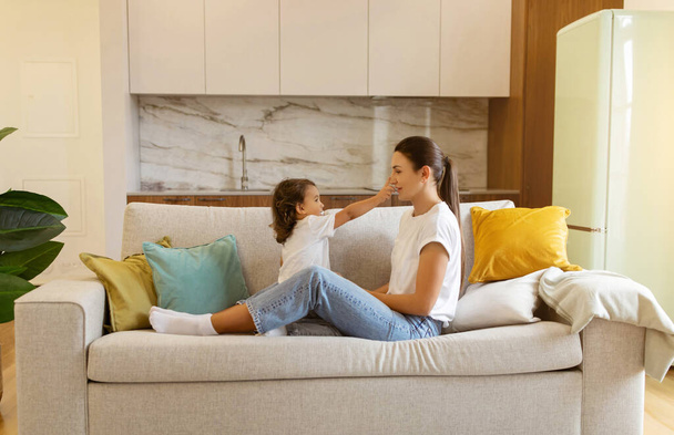 Young mother and her preschool daughter enjoying playtime on sofa at home, happy mom and cute little female child having fun together in living room, symbolizing loving family bonds, copy space - Foto, Imagen