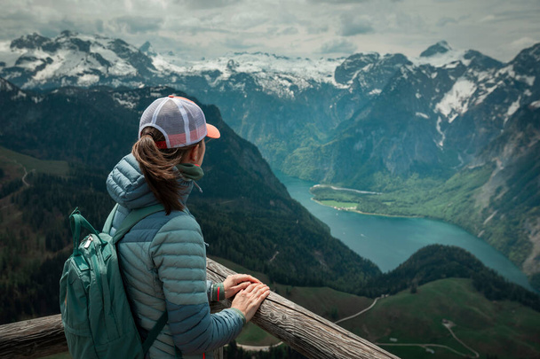 Female hiker with backpack looking at turquoise lake Knigssee from above viewpoint of mountain peak Jenner at Berchtesgaden Bavaria, snow-capped mountains in the background - Fotografie, Obrázek