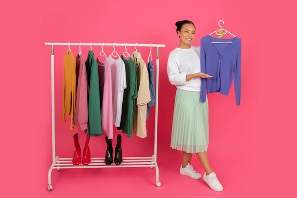 Happy asian female showing new blouse while standing near clothing rack, smiling korean shopper lady exploring different styles, posing isolated on pink studio background, full length shot - Zdjęcie, obraz
