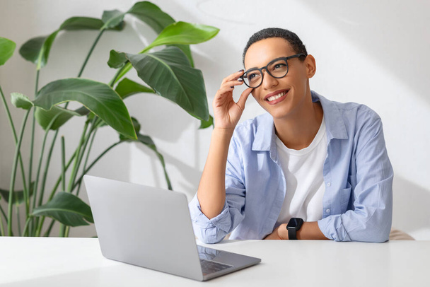 Positive millennial latin lady in glasses at table with computer, enjoy work and study, in white cafe, office interior. Communication remotely, app, gadget for business, freelance - 写真・画像