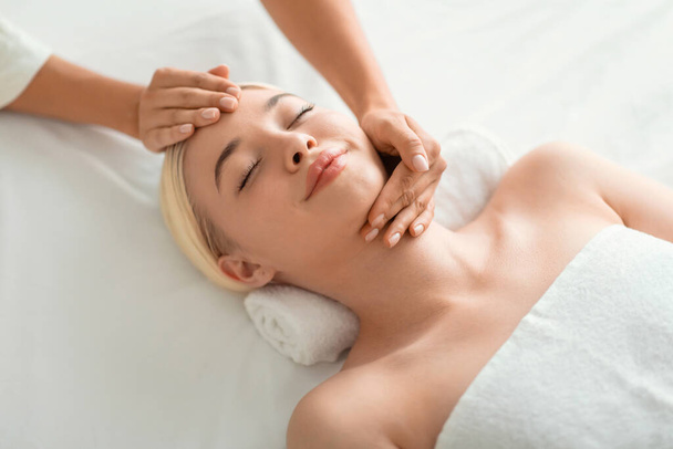 Beauty And Relaxation. Young blonde woman enjoying facial massage, lying on spa bed indoor at beauty salon, high angle shot of lady relaxing during facebuilding treatment - Foto, immagini