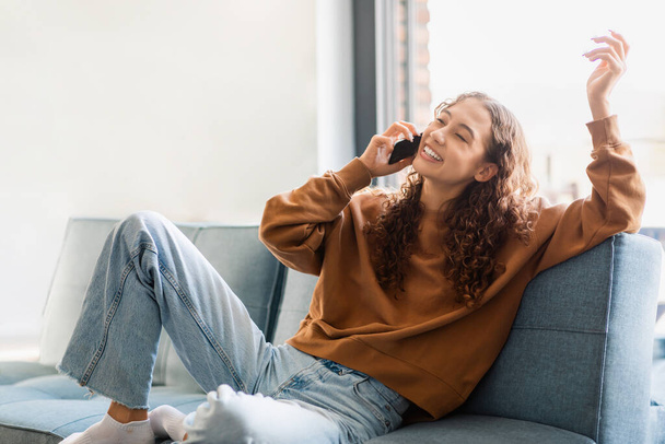 Teen girl chatting on phone comfortably lounging on couch at home with favorite device, adolescent lady deeply engrossed in pleasant digital conversation with friend, smile signifying happy news - Foto, Imagem