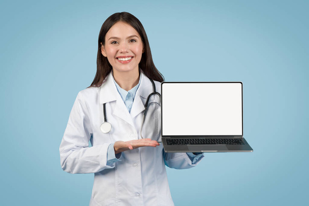 Smiling female doctor presenting laptop computer with blank screen for customizable content, ideal for telehealth ads, on blue background, mockup - Foto, Bild