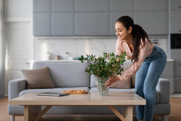 Smiling woman in peach blouse and blue jeans carefully arranges plant in vase on wooden coffee table in bright, modern living room setting - Foto, Bild