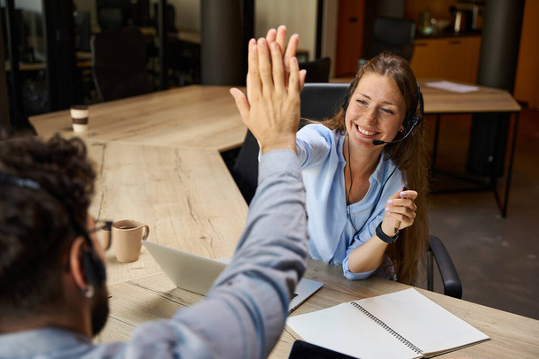 Young smiling caucasian call center operators giving high five each other during work at table in coworking office. Concept of teamwork - Foto, Bild
