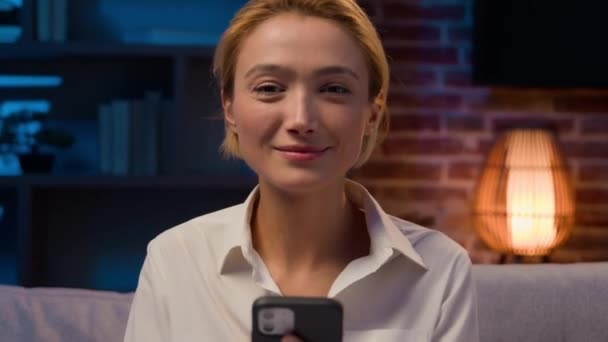 Кавказская беззаботная женщина с мобильным телефоном улыбается в камеру ночью дома на диване счастливая деловая женщина в вечернем офисе со смартфоном сотового телефона социальные медиа удовлетворены интернет-службы улыбка - Кадры, видео