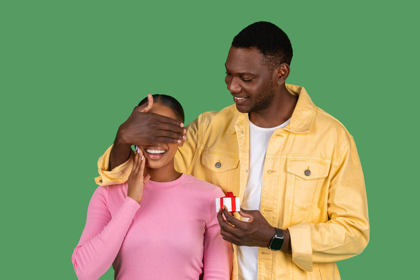 Black guy make anniversary surprise to his girlfriend, young man close woman eyes, holding little gift box with jewelry engagement ring, green background. Proposal, Valentine Day celebration - Foto, Bild