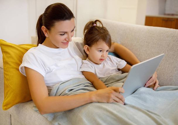 Mom And Little Daughter Lying Under Blanket And Using Digital Tablet Together, Happy Young Mother And Her Toddler Female Child Relaxing With Modern Gadget At Home, Enjoying Online Entertainment - Фото, зображення